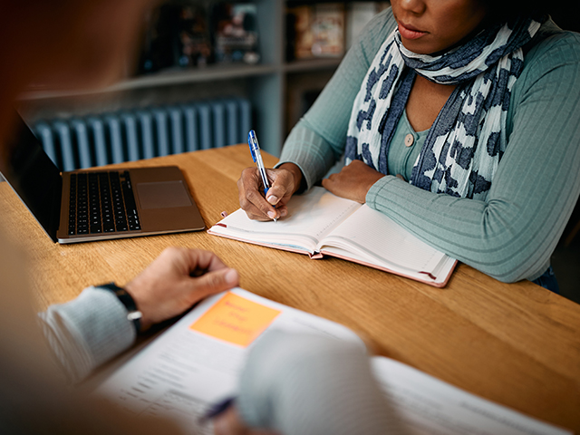 Image of a woman seated at a table, writing in a notebook with a pen during a tutoring session