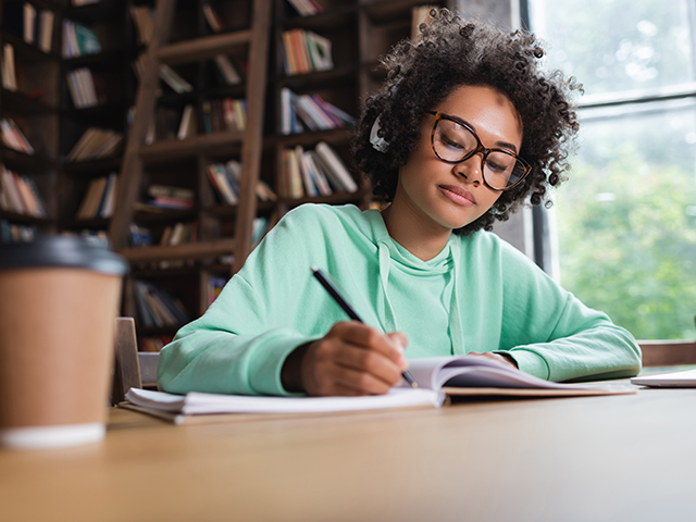 girl in green hoodie writing in notebook at desk