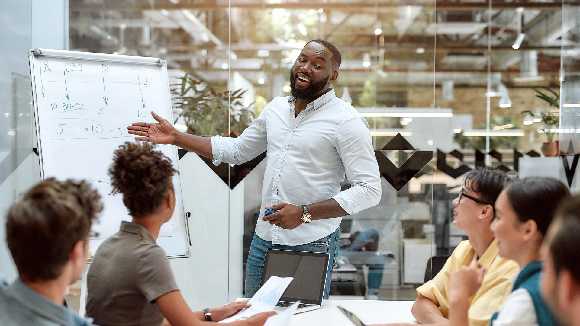 Young african american male student doing a presentation to team group during internship, pointing at whiteboard and smiling