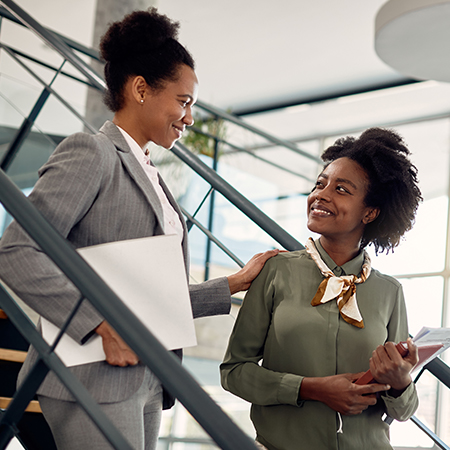 Happy black student talks to female businesswoman mentor who leads her through office building for internship.