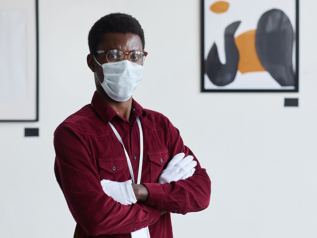 Waist up shot of African-American man wearing mask looking at camera while standing with arms crossed against modern art paintings in gallery