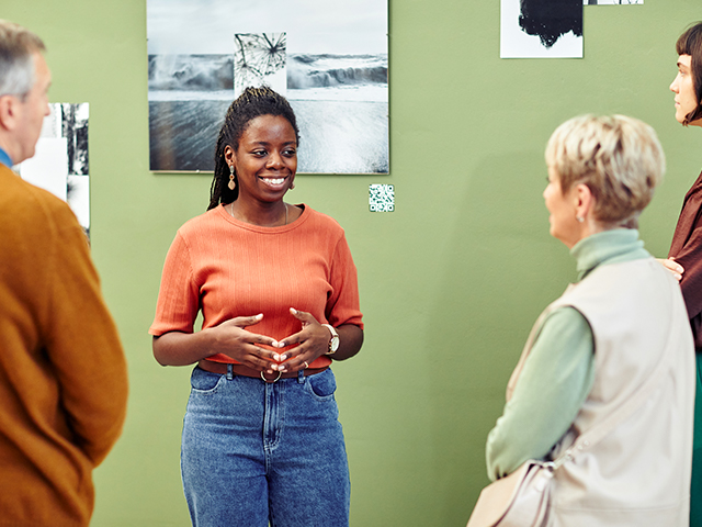 Cheerful young black female creator standing in front of art gallery exhibition visitors speaking about art 